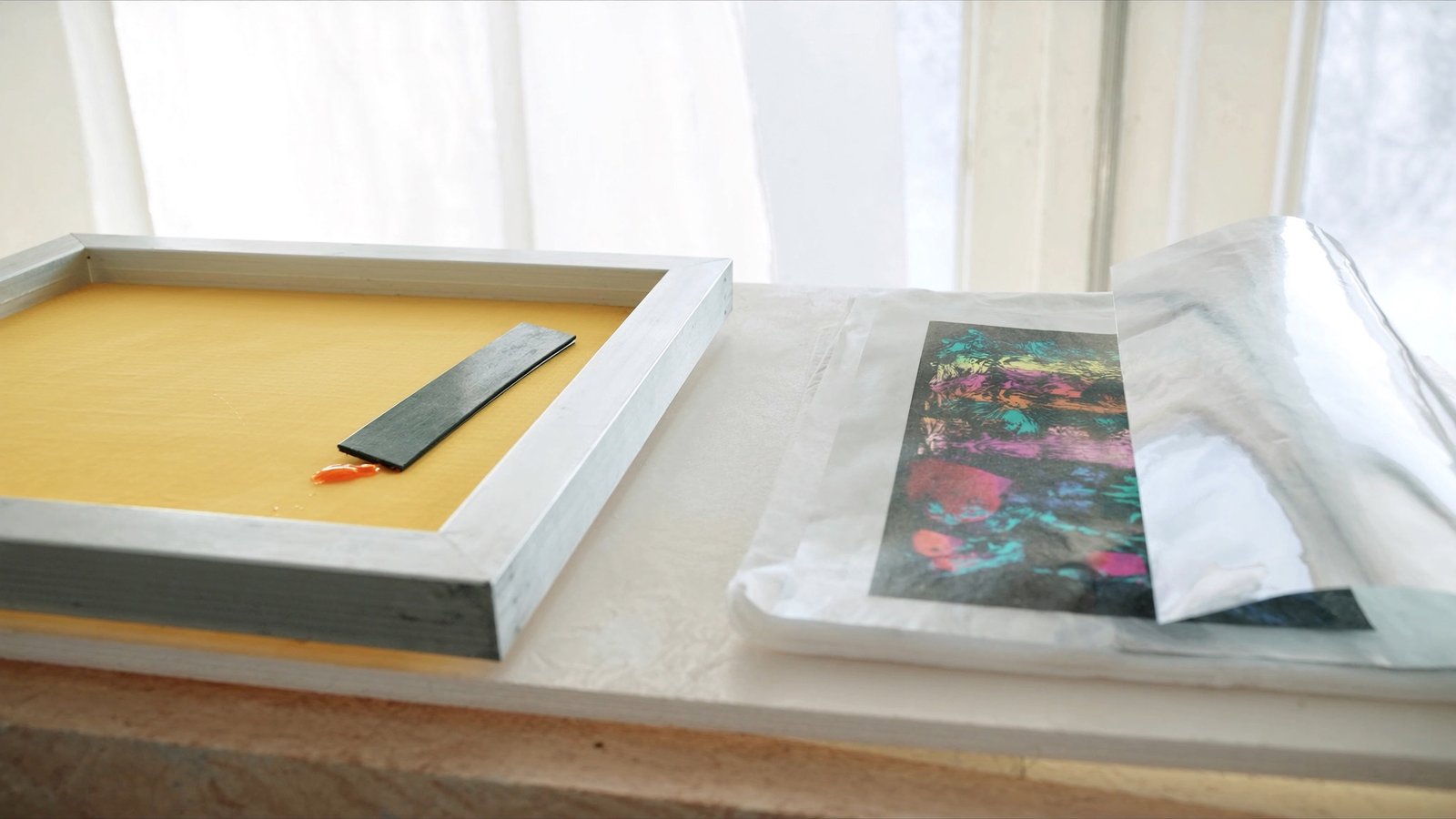 Telaio serigrafico con maglia gialla, lama della racla e inchiostro arancione accanto a una pellicola DTF lucida stampata a colori, alla luce di una finestra